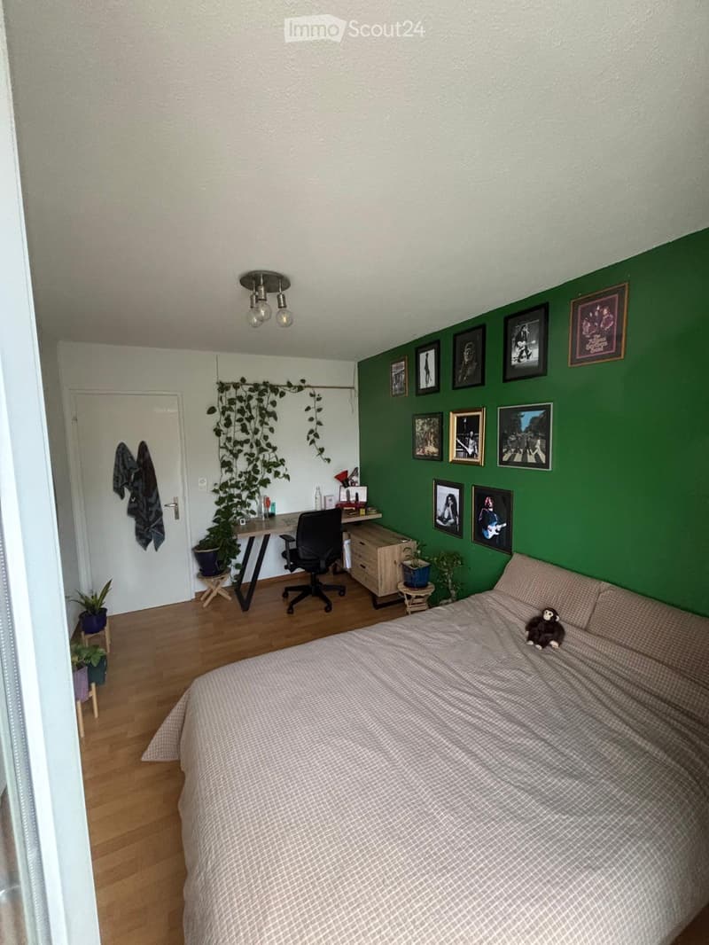 Cozy bedroom with a bed, desk, chair, and several framed pictures on the wall. Wooden floor and green accent wall.