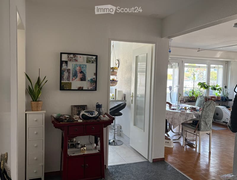 A well-lit hallway with a small table and stool on the left, a cabinet with drawers, and a doorway leading to a dining area with a table, chairs, and plants. The walls are adorned with picture frames and a potted plant is placed on a small cabinet.