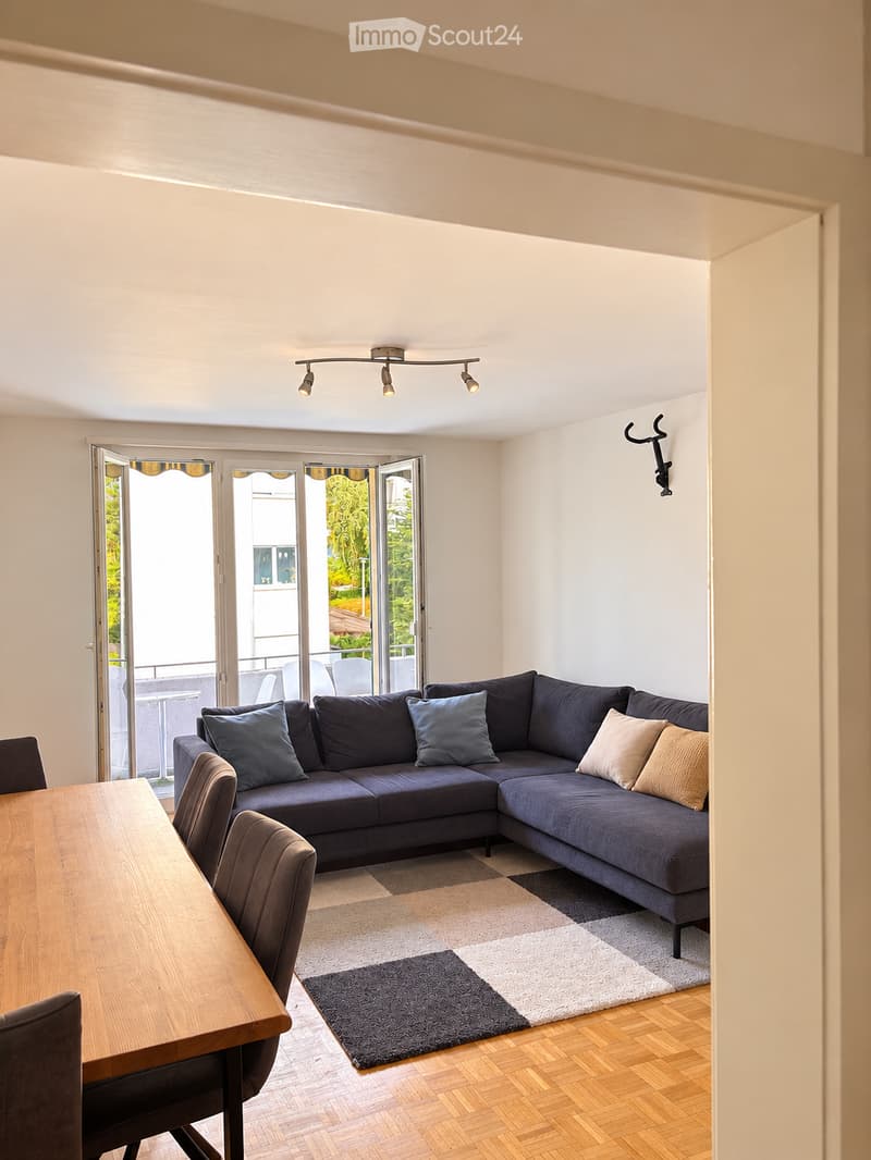 Modern living room with a grey sectional sofa, wooden dining table, chairs, and a patterned rug. Large sliding glass doors open to a balcony with outdoor seating.
