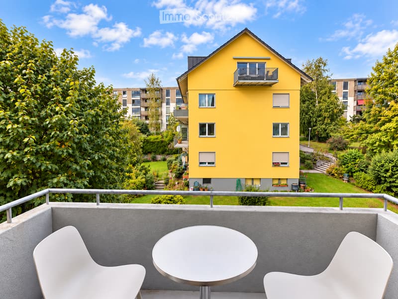 Balcony with a round table and two white chairs, overlooking a garden and a yellow house with multiple windows and balconies.