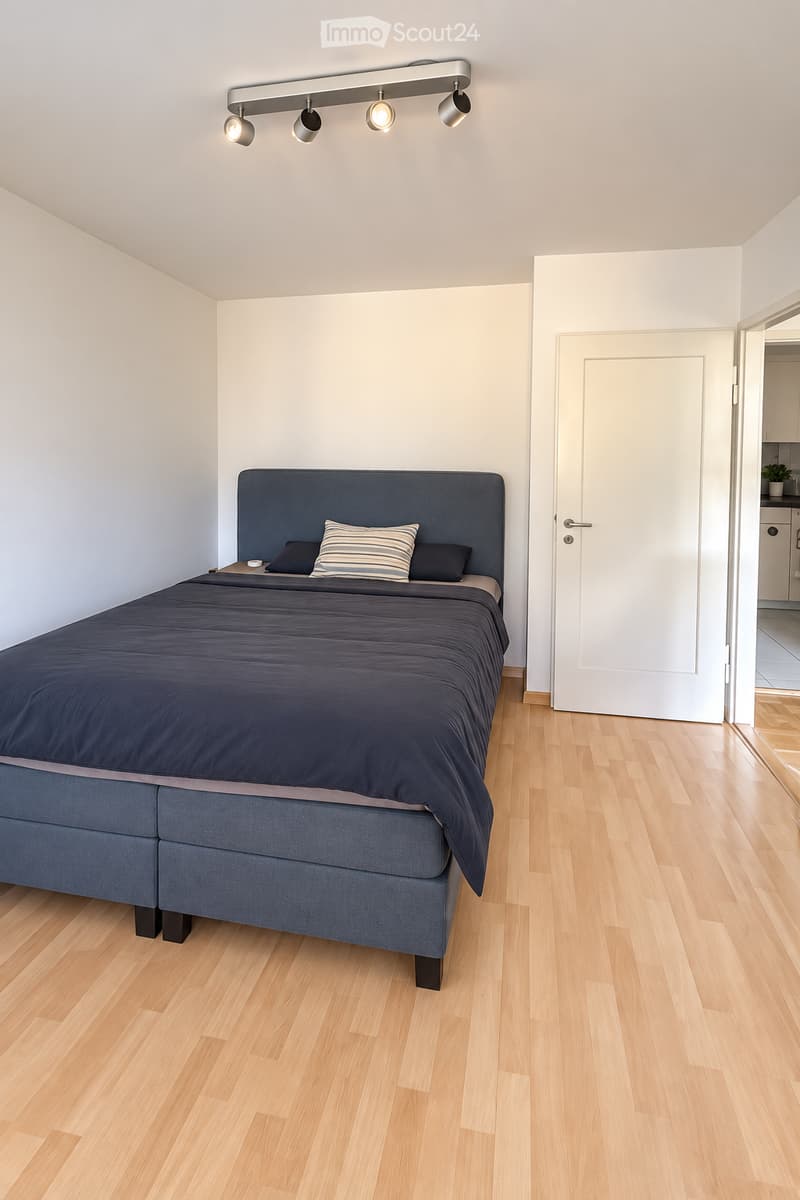 Modern bedroom with a large bed, dark blue bedding, and a nightstand. The room features light wooden flooring and white walls, with a door leading to another room visible in the background.