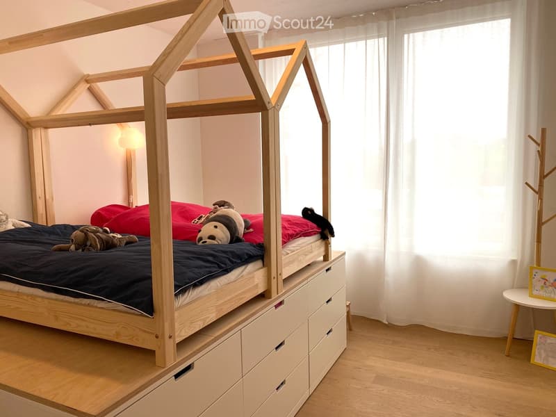 A modern bedroom with a lofted wooden bed, red pillows, stuffed toys, a small table with picture frames, and a coat rack.