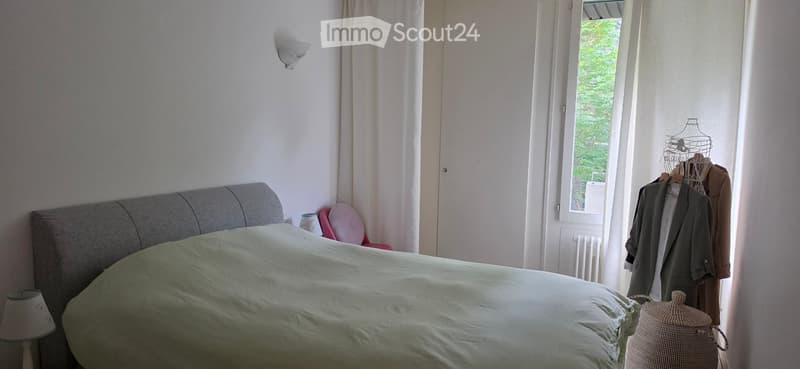 Cozy bedroom with a grey headboard, white bedding, nightstand, lamp, and a chair near the window. Clothes are hanging on a rack near the window.