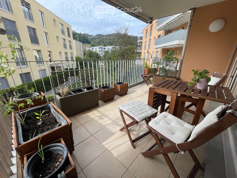 Balcony with wooden table, chairs, potted plants, and a view of the surrounding buildings and greenery.