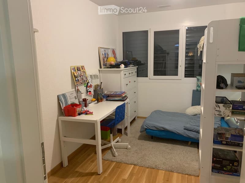 A compact bedroom with a single bed, a desk with a chair, a cabinet, and various personal items including books, a ball, and storage boxes.