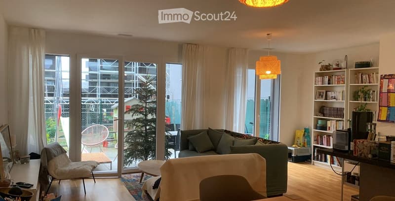 Bright living room with large windows and sliding glass doors leading to a balcony. Features a green sofa, a white chair, a small table, a bookshelf, and a hanging lamp.