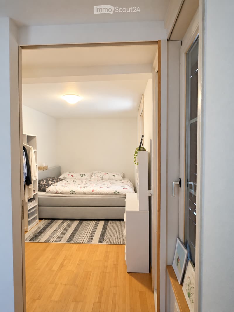 Cozy, compact bedroom with wooden flooring, a bed with floral bedding, a nightstand with a mirror, a small plant, and a wardrobe with drawers. A patterned rug lies in front of the bed.