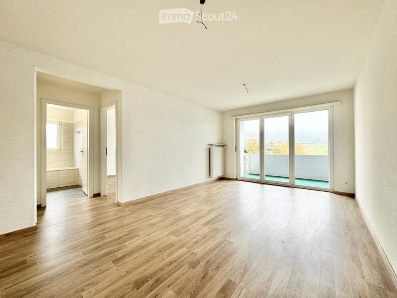 Empty room with wooden flooring, white walls, large sliding glass doors leading to a balcony, and an adjacent bathroom visible through an open doorway.