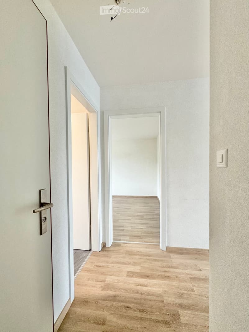 Empty hallway with light wooden flooring, white walls, and two open doors leading to other rooms.