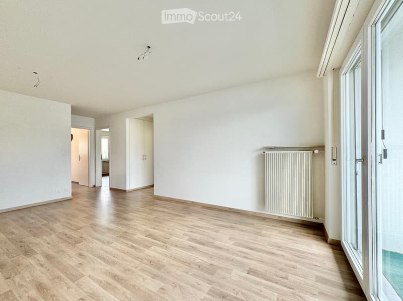 Empty room with light wooden flooring, white walls, and a radiator. Features a large glass door leading outside and several open doorways to adjacent spaces.
