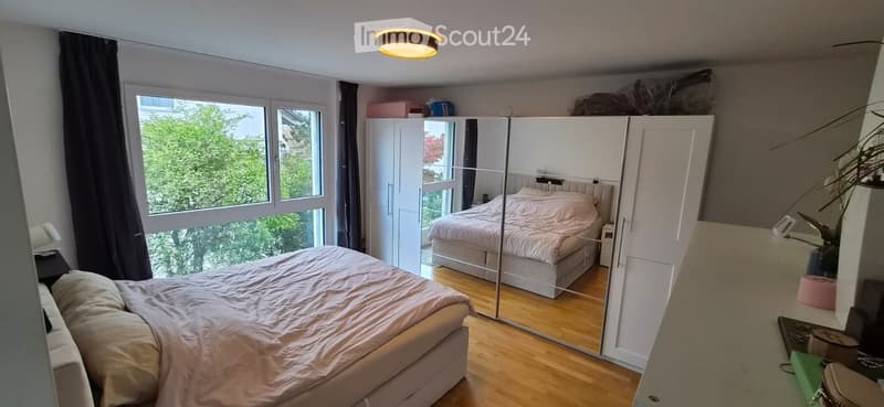 Bright bedroom with wooden flooring, large window with view, double bed with beige bedding, mirrored wardrobe, nightstand, and lamp.