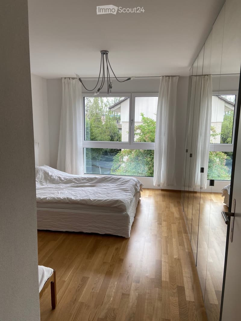 Bright bedroom with wooden flooring, large window with white curtains, and a neatly made bed.