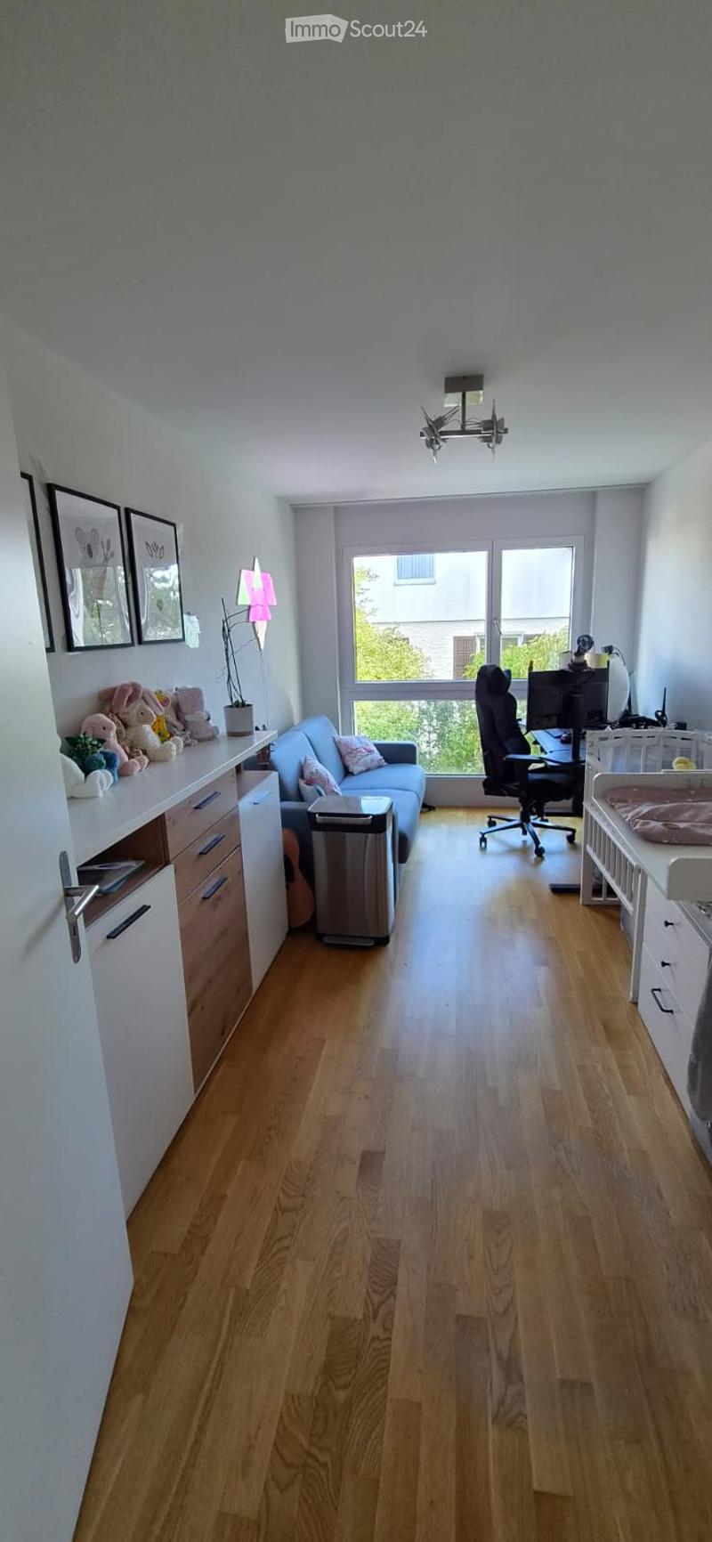 A multi-functional room with a light blue sofa, a black office chair, a desk, a small storage unit, and a baby crib. The room features wooden flooring, large windows, and framed pictures on the wall.