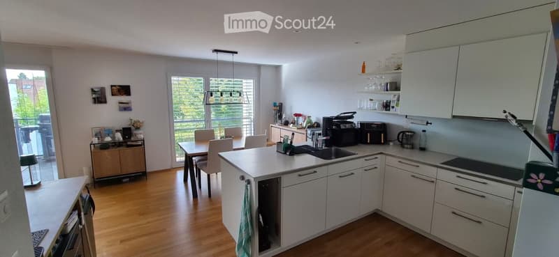 Modern kitchen with white cabinets, a central island, and a dining area. The space features a sink, coffee maker, toaster, microwave, and various kitchen appliances. There is a dining table with chairs and a lamp on the sideboard.