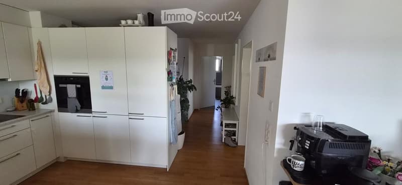 A well-lit hallway with wooden flooring, leading to a kitchen area with white cabinets, a sink, and various kitchen utensils. There is a coffee machine on a small table with a mug and a vase of flowers nearby.