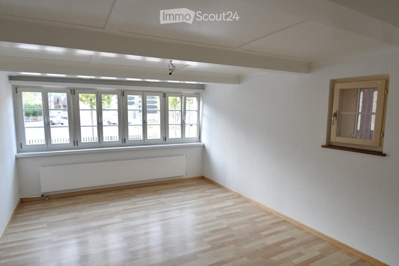 Empty room with wooden flooring, white walls, large windows, radiator, and a small wooden shelf on the right side.