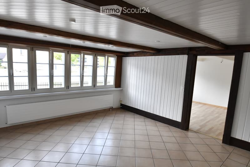 Empty room with exposed wooden beams, tiled floor, large windows, radiator, and white paneled walls.