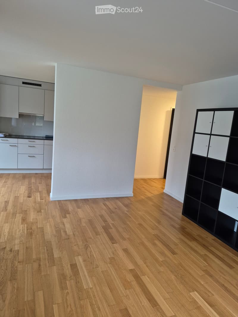 Empty room with wooden flooring, white walls, and a partially visible kitchen area with white cabinets and drawers.