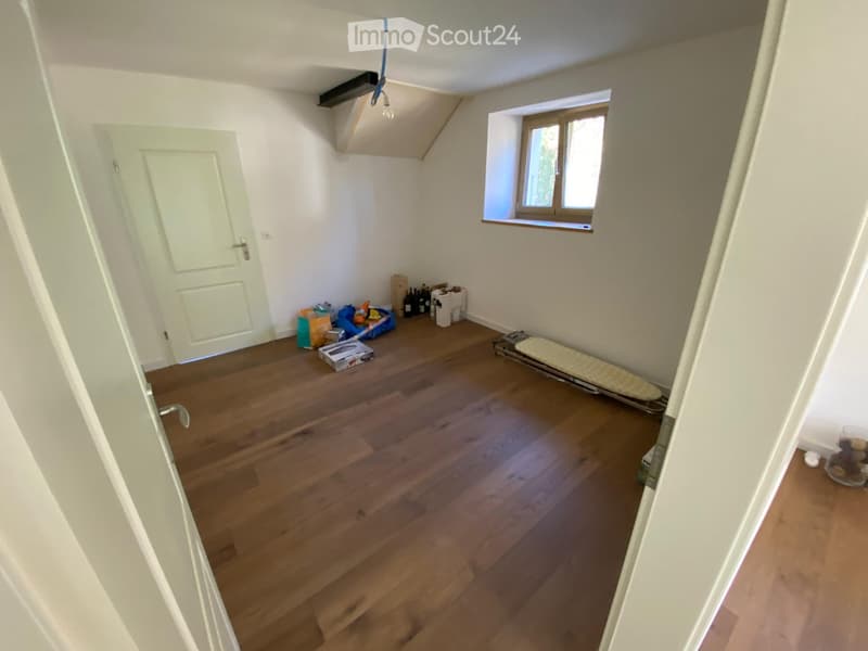 Empty room with wooden flooring, white walls, a window, and a door. Some storage items are visible near the window.