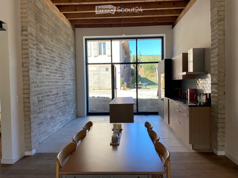 Bright kitchen with exposed brick walls, large glass doors leading to an outdoor area, modern cabinets, and a central island with a dining table and chairs.
