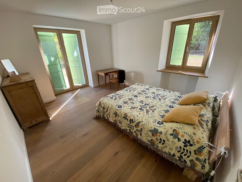 Bedroom with wooden flooring, a bed with a colorful patterned bedspread, a small table and chair, and a large window with a view of greenery.