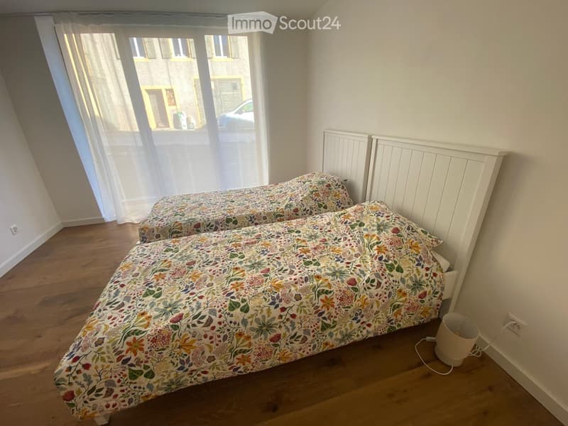 Cozy bedroom with two single beds, floral bedding, wooden flooring, radiator, and large window with white curtains.