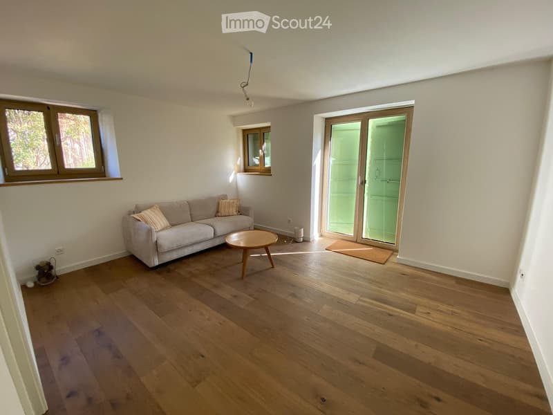 Bright living room with wooden flooring, a grey sofa with throw pillows, a round wooden coffee table, and large windows with a sliding glass door leading outside.