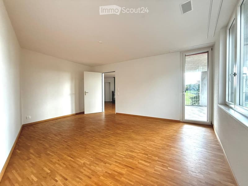 Empty room with wooden flooring, white walls, and large windows. Features a sliding glass door leading outside and an open doorway to another room.