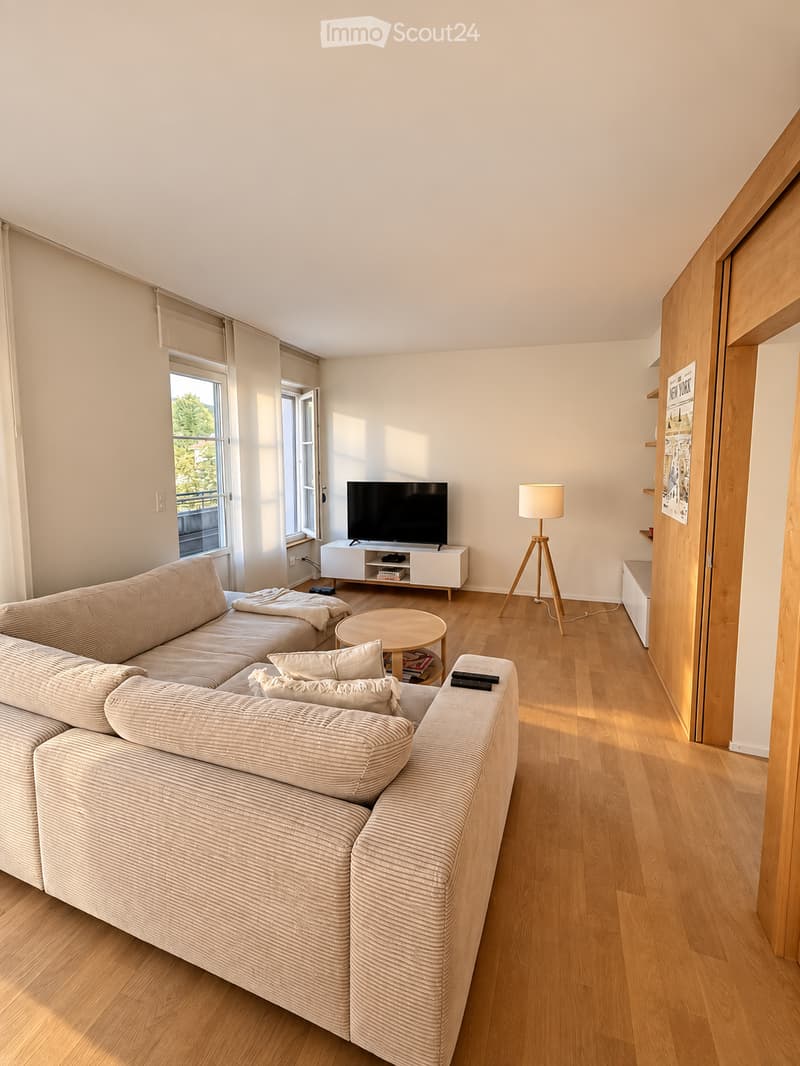 Modern living room with light wooden flooring, large windows, a sectional sofa, a coffee table, a television on a white stand, and a floor lamp.