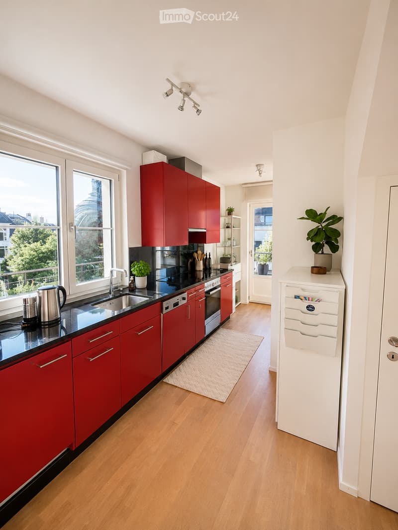 Modern kitchen with red cabinets, black countertops, stainless steel appliances, and wooden flooring. Features include a sink, oven, dishwasher, refrigerator, and various kitchen items on the counter.
