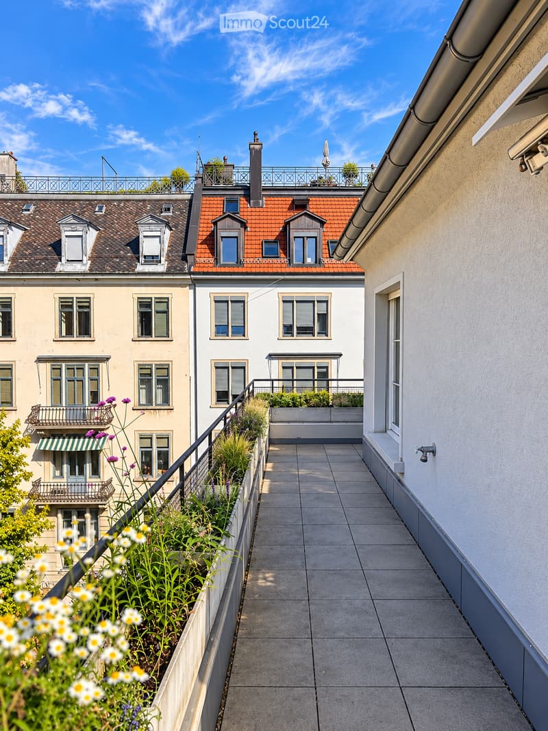 Balcony with tiled floor, metal railing, and potted plants, overlooking neighboring buildings with red-tiled roofs and multiple windows.