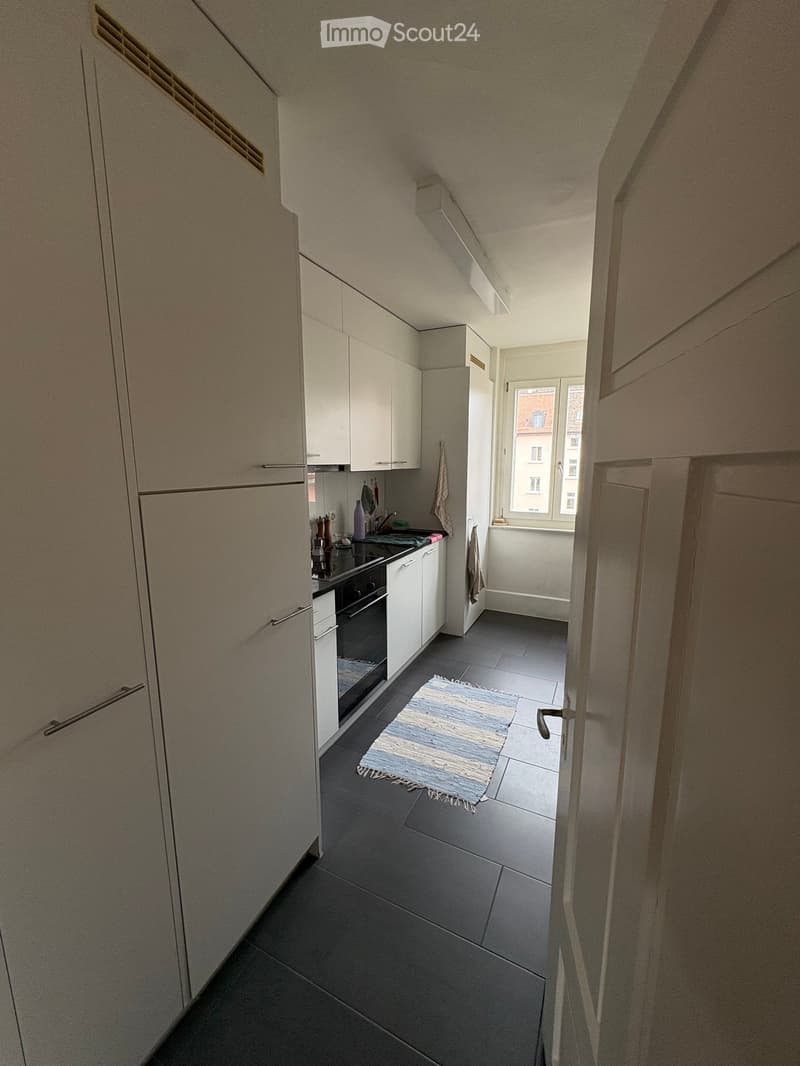 Modern kitchen with white cabinets, dark tiled floor, and a small rug in the center. Features an oven, faucet, and various kitchen items on the counter.