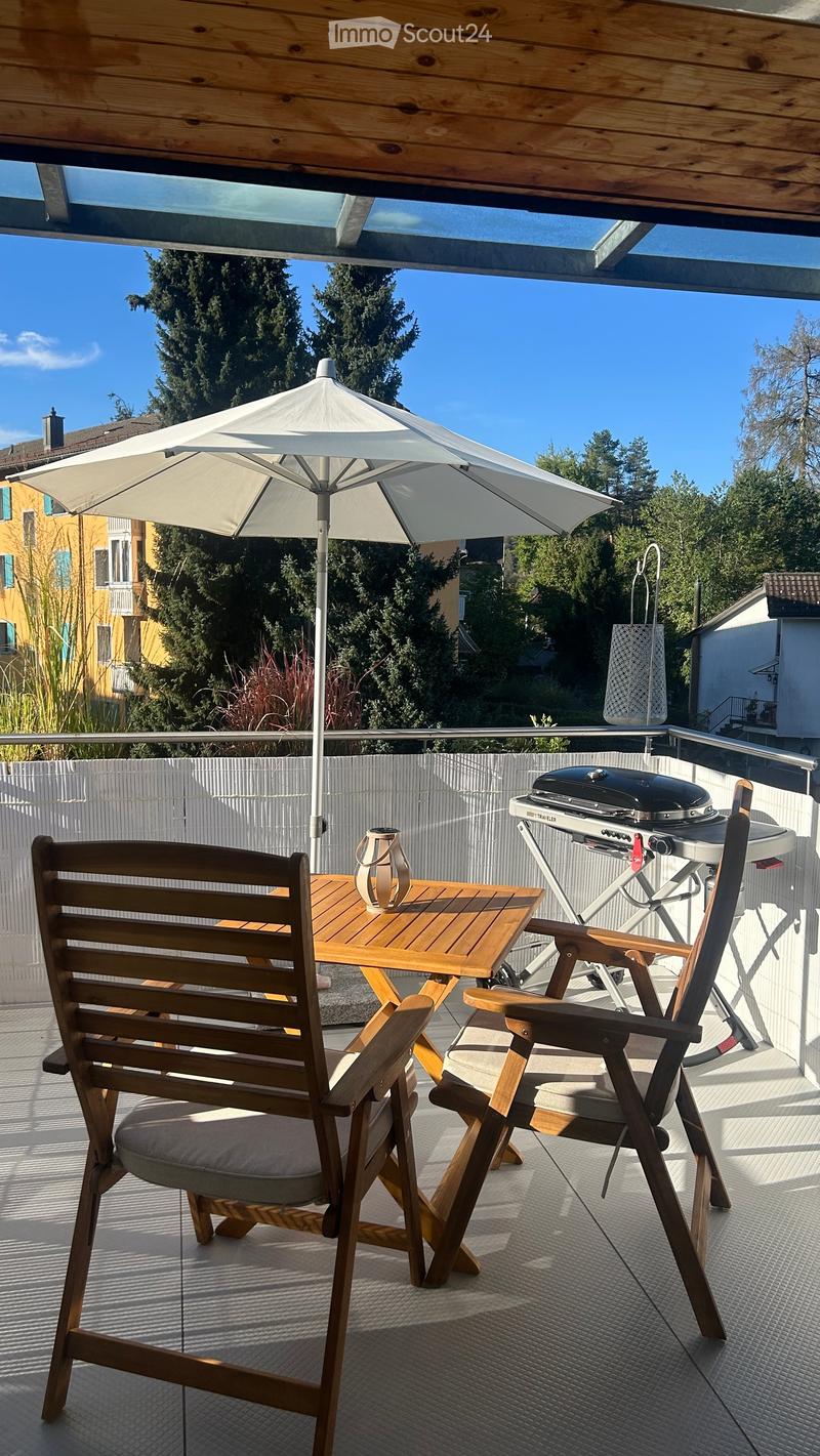 Balcony with wooden table, two chairs, umbrella, and grill, surrounded by trees and a clear blue sky.