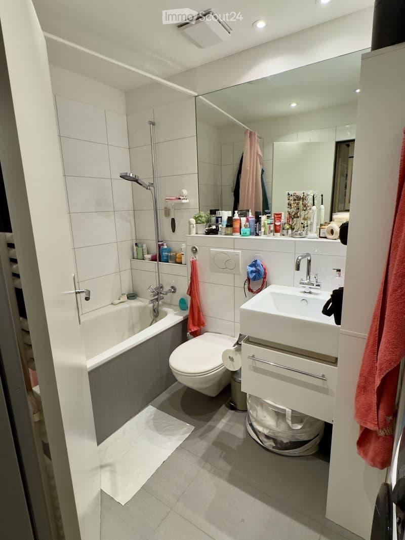 Modern bathroom with white tiles, bathtub, toilet, sink, large mirror, and various toiletries on the counter.