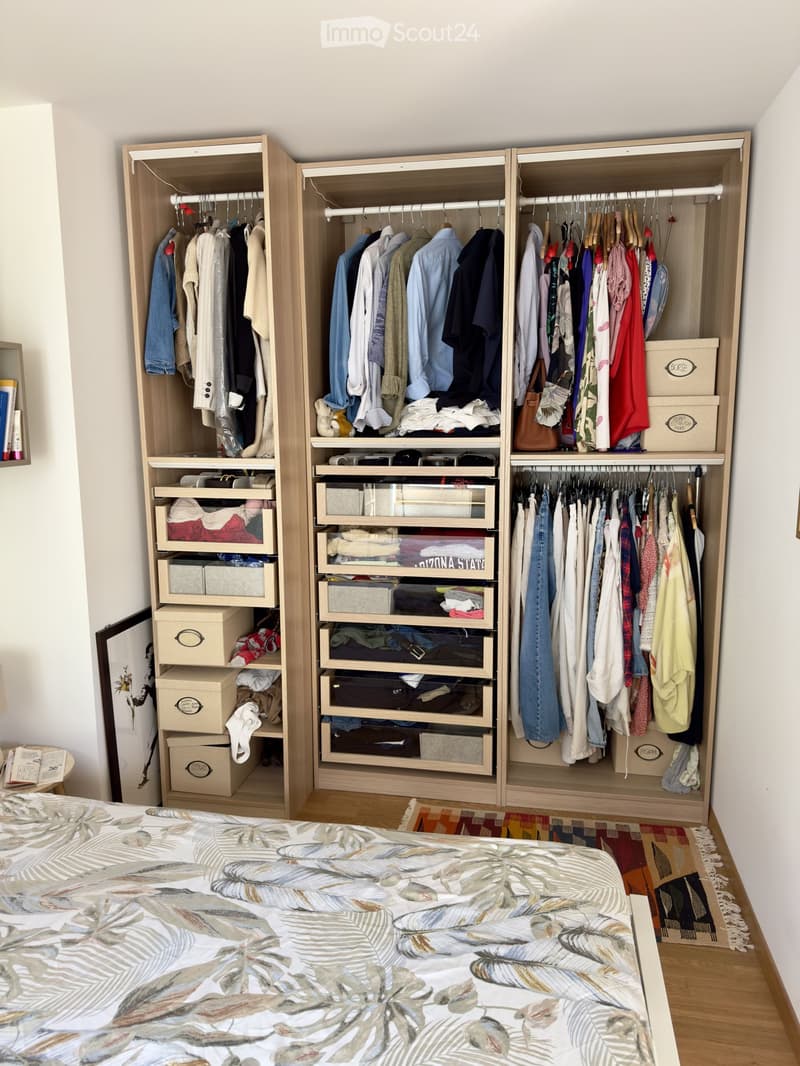 A well-organized bedroom with a large bed, a tall wooden wardrobe filled with clothes and accessories, multiple storage drawers, and a small table with books.