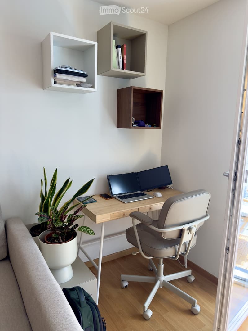 A modern, minimalist workspace with a wooden desk, laptop, ergonomic chair, potted plants, and built-in wall shelves holding books and other items.