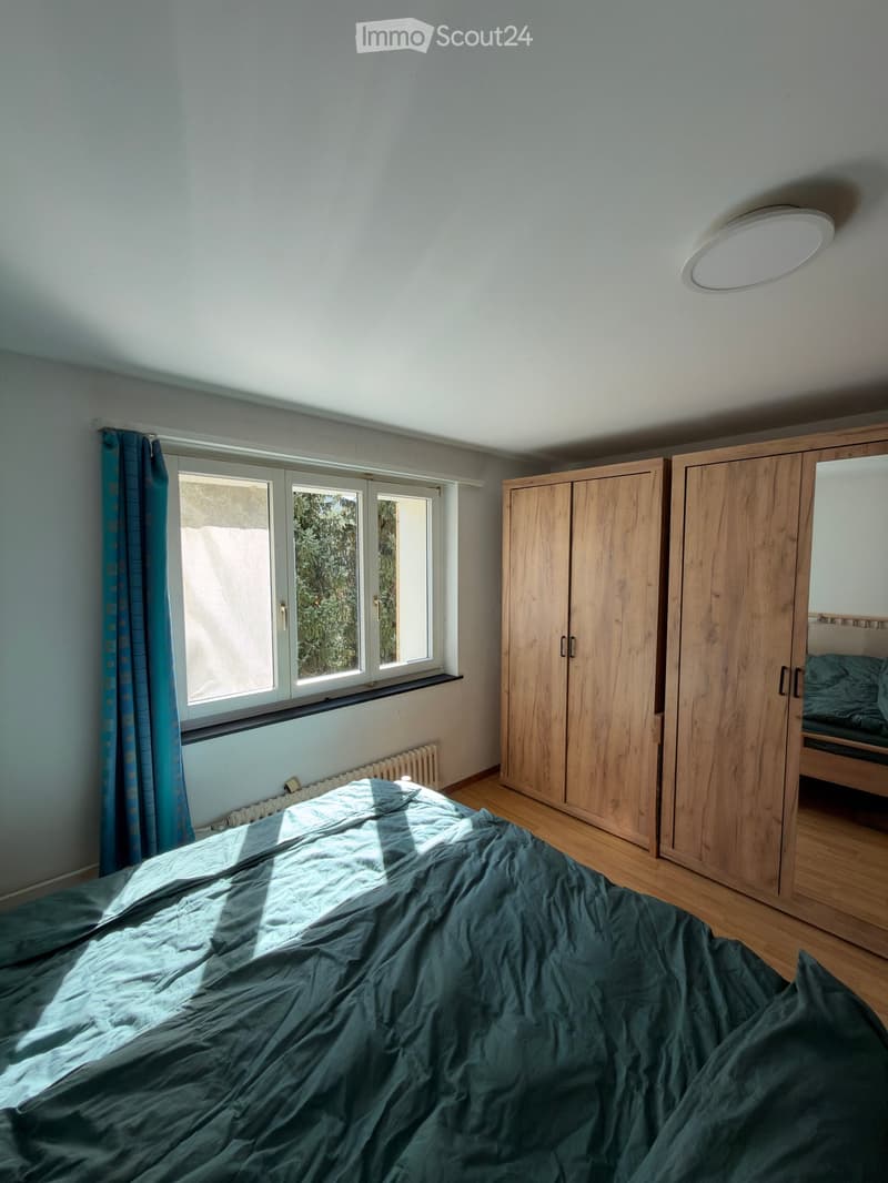 Bright bedroom with wooden floor, large window with blue curtain, bed with dark green duvet, wooden wardrobe with mirror, and a round ceiling light.