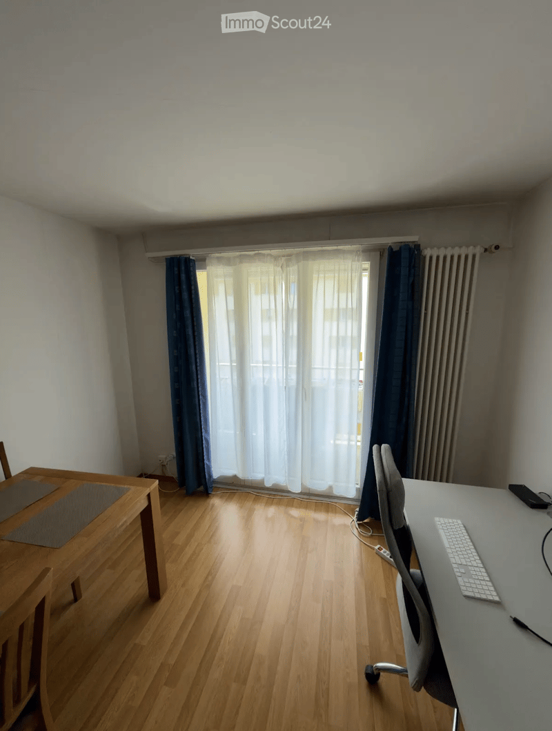 A small, unfurnished room with wooden flooring, a desk with a chair, a small dining table with chairs, and a window with curtains.