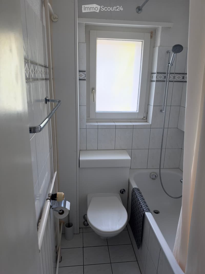 Modern bathroom with white tiles, a toilet, bathtub, shower, towel, and toilet paper holder.