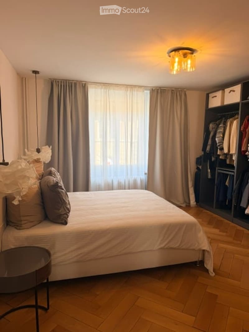 Modern bedroom with a large bed, nightstand, and built-in wardrobe. Natural light filters through large windows with curtains. Wooden flooring and contemporary lighting.