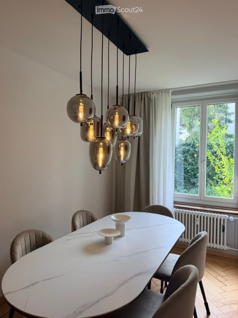 Modern dining room with a white marble dining table, six chairs, and a stylish pendant light fixture above. Large window with curtains and radiator visible.