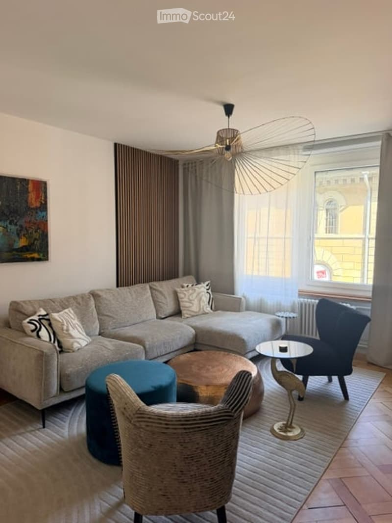 Modern living room with a grey sectional sofa, coffee table, blue and beige chairs, a small round table, and a decorative lamp. Large windows with curtains allow natural light to fill the space.