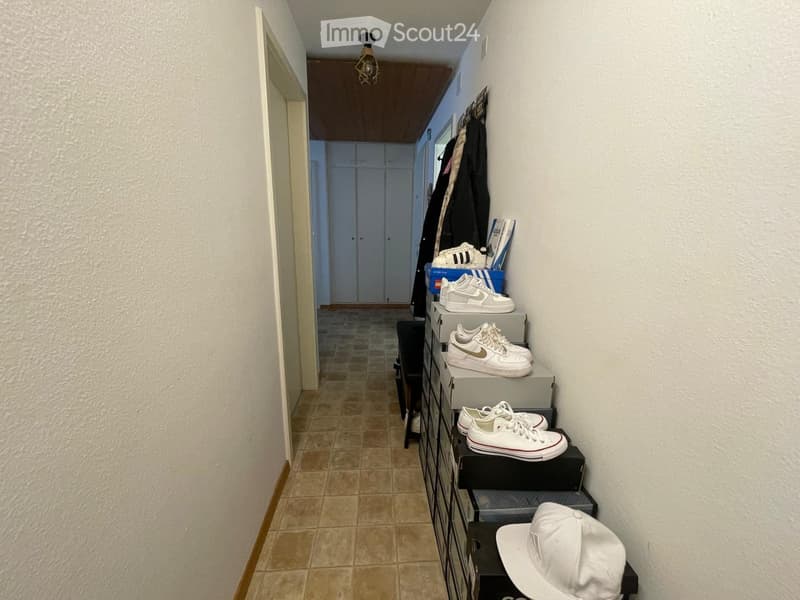 A narrow hallway with tiled flooring, white walls, and a ceiling light fixture. A small shelf on the right holds several pairs of white sneakers, a white baseball cap, and some boxes.
