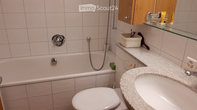 Bathroom with a bathtub, toilet, sink, mirror, and wooden cabinet. Includes a wicker basket and various toiletries on the countertop.