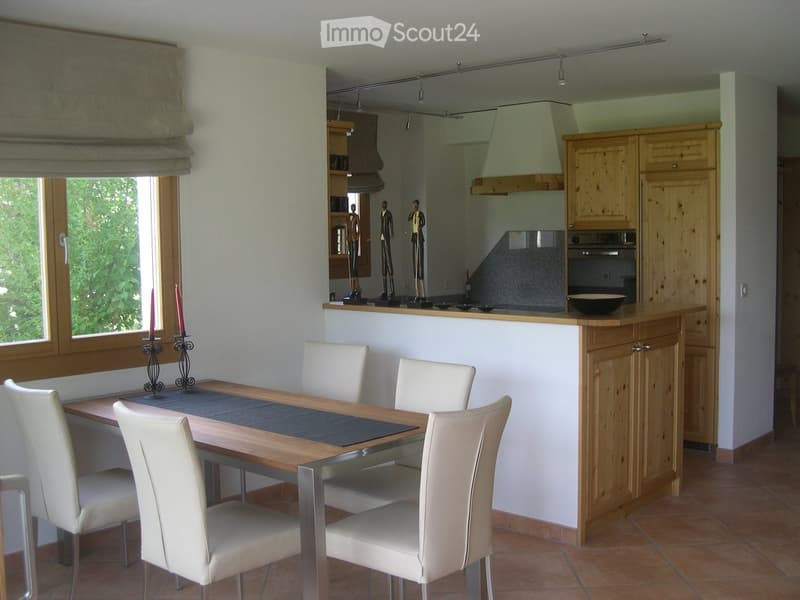Bright kitchen and dining area with wooden cabinets, a dining table with white chairs, and a window with blinds. The kitchen features an oven, a bowl on the counter, and decorative figurines on a shelf.