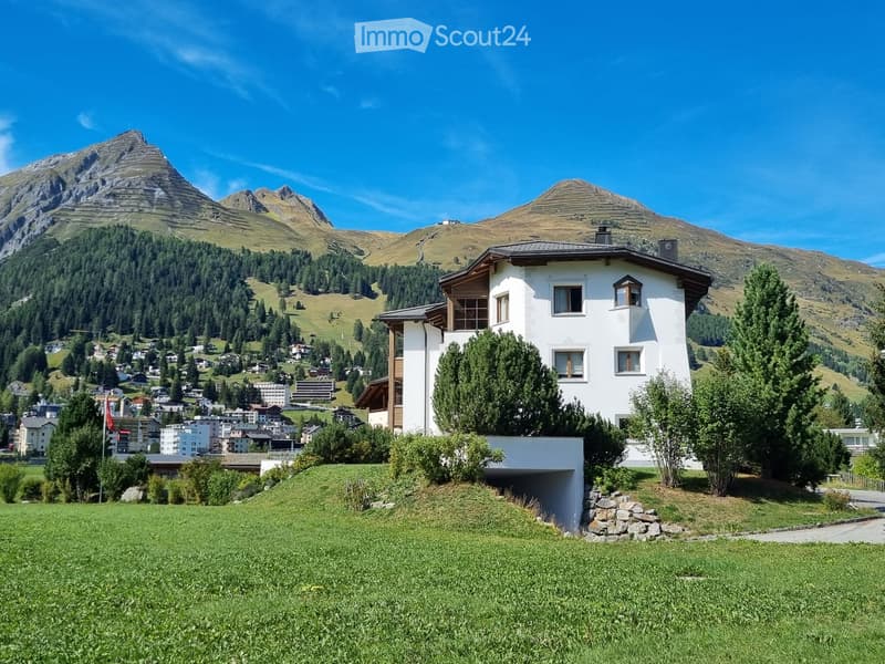 A modern white house with a sloped roof and multiple windows, situated on a grassy slope with a panoramic mountain view in the background.