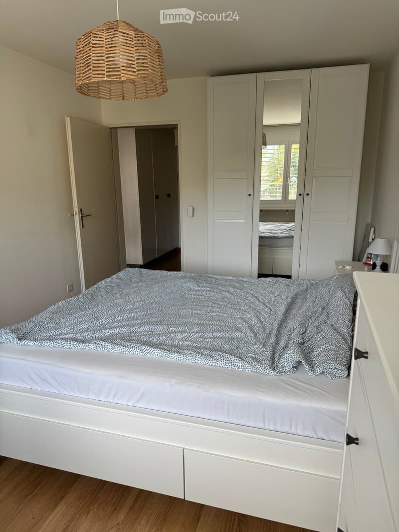 A modern bedroom with a white bed, a large wardrobe, and a hanging woven lamp. The room features wooden flooring and an open doorway leading to another room.