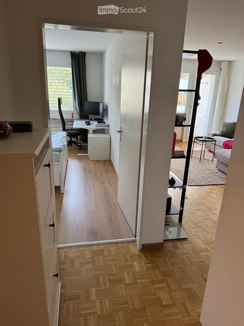 A modern, open-plan living and working space with wooden flooring, large windows, and a mix of furniture including a sofa, desk, and chair.