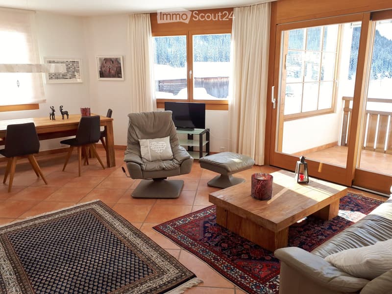 A well-furnished living room with a sofa, coffee table, armchair, TV, and a dining area with a table and chairs. Large windows offer a scenic view, and the space is decorated with rugs and artwork.