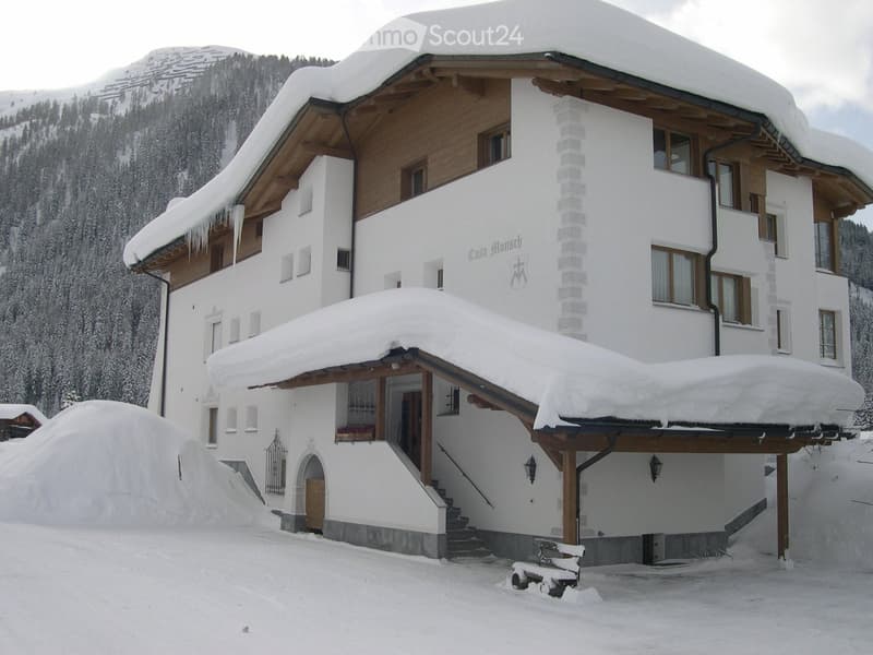 A large, white, multi-story house with a snow-covered roof and wooden accents, situated in a snowy mountain landscape.